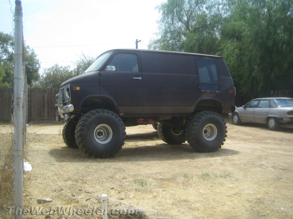 Dark-colored van on massive off-road tires, parked next to a sedan