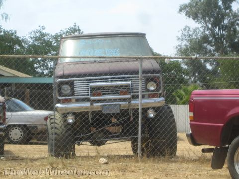 1968 Ford Mustang - Front view of a maroon/burgundy lifted Ford van (early-1970s Econoline style) sitting on oversized mud tires behind a chain-link fence, wit…