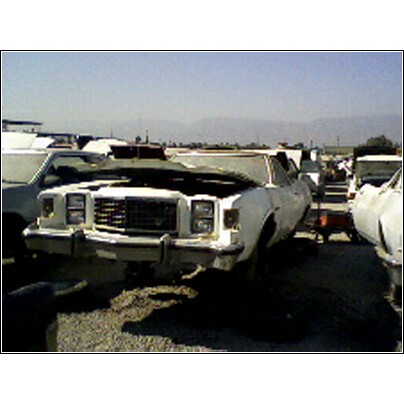 1973 Ford Ranchero - A white sedan sits in a junkyard with its hood open, surrounded by other vehicles under a clear sky.