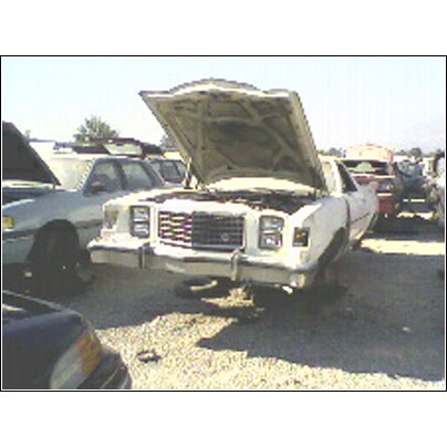 1973 Ford Ranchero - A white car sits in a junkyard with its hood open, showing the front grille and headlights.