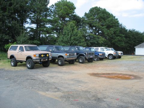 1995 Ford C-350 (Centurion Conversions) - A row of lifted Ford trucks and Broncos are parked in a lot, with trees and a building in the background.