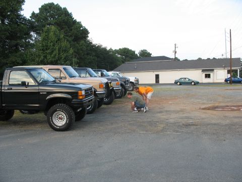 1995 Ford C-350 (Centurion Conversions) - A row of pickup trucks is parked in a lot, with two men inspecting the ground near the front vehicles.