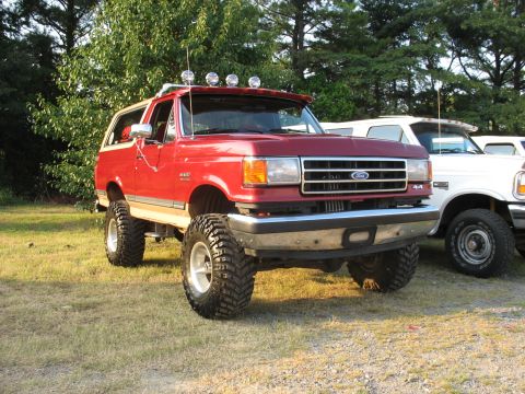 1995 Ford C-350 (Centurion Conversions) - A lifted red Ford Bronco with large tires and a light bar sits in a grassy area next to other vehicles.