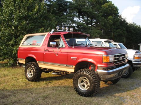 1995 Ford C-350 (Centurion Conversions) - Modified red Ford Bronco with a tan roof and large off-road tires parked outdoors next to other vehicles.