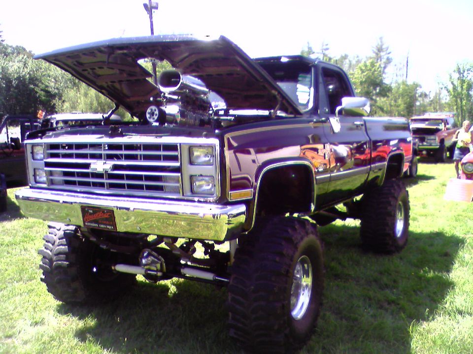 Lifted dark square-body Chevy/GMC pickup with hood up showing blown engine, on big mud tires.
