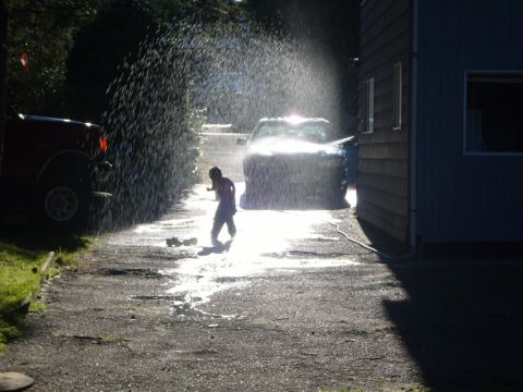 1989 Ford Bronco - A child plays with a hose, spraying a dark SUV in a driveway next to a red truck.