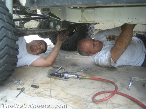 1987 Suzuki Samurai - Mechanics are performing maintenance on a vehicle's undercarriage, surrounded by tools and equipment.