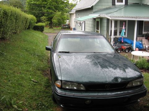 1992 Ford Crown Victoria - A dark green sedan sits on a wet driveway, with a house and yard visible in the background.