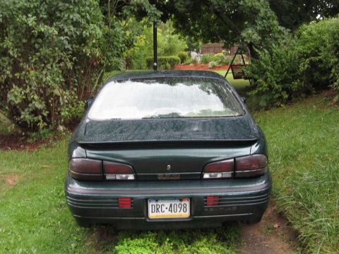 1992 Ford Crown Victoria - The rear of a green sedan is shown parked in a grassy yard with trees and a swing set visible.