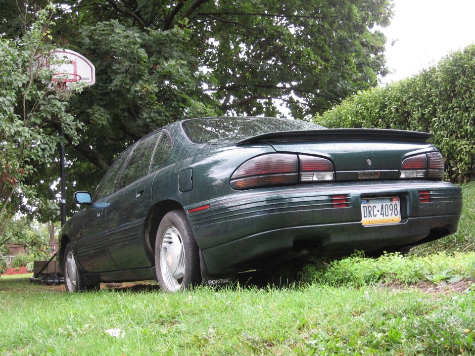 Green sedan parked on grass, rear view showing taillights, spoiler, and license plate DRC-4098