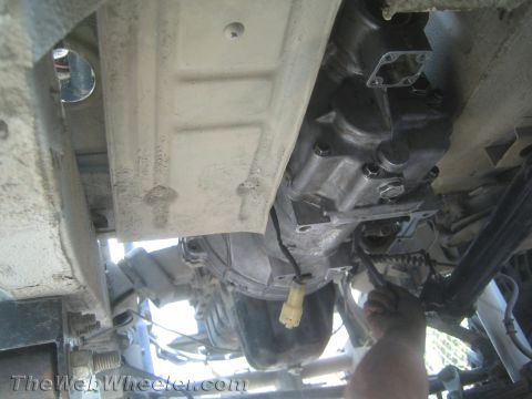 1987 Suzuki Samurai - A close-up view of a vehicle's transmission and undercarriage, showing mechanical components and a hand performing work.