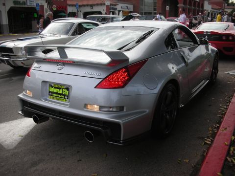 1998 Ford Crown Victoria - A silver Nissan 350Z Nismo is parked on the street, showing its distinctive rear design and NISMO badging. Other vehicles are visible in th…