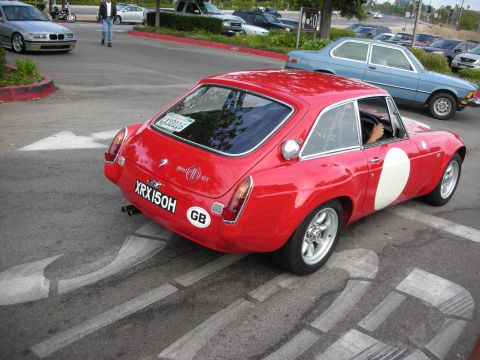 1998 Ford Crown Victoria - A red MGB GT with a white racing stripe is parked on a street, with other vehicles visible in the background.