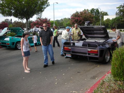 1998 Ford Crown Victoria - Several people gather around a purple sports car with its rear hood open at an outdoor event.