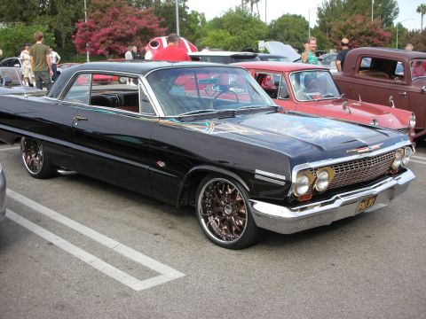 1998 Ford Crown Victoria - A black 1963 Chevrolet Impala with custom chrome wheels and intricate hood artwork is parked at a car meet.