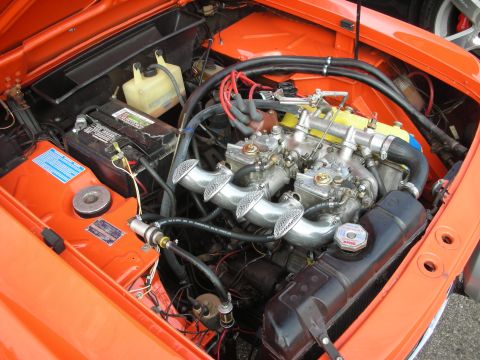 1998 Ford Crown Victoria - The engine bay of an orange car is shown, revealing its V6 engine with dual carburetors and a battery.