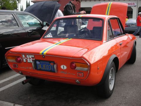 1998 Ford Crown Victoria - An orange Lancia Fulvia HF coupe with racing stripes is parked next to another car, with its hood open.