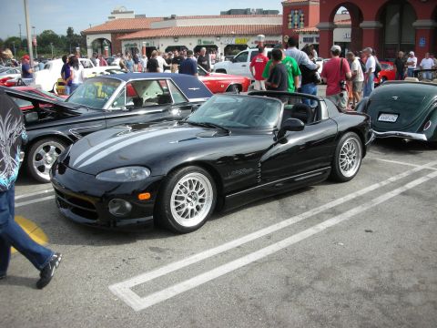 1998 Ford Crown Victoria - A black convertible sports car with silver racing stripes is parked at a car meet, alongside other vehicles.