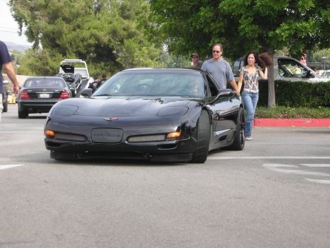 1998 Ford Crown Victoria - A black Chevrolet Corvette C5 is seen from the front, with two people standing next to it. Another car is visible in the background.