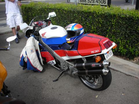 1998 Ford Crown Victoria - A Honda RC30 motorcycle is parked on the street next to a hedge, with a person standing nearby.