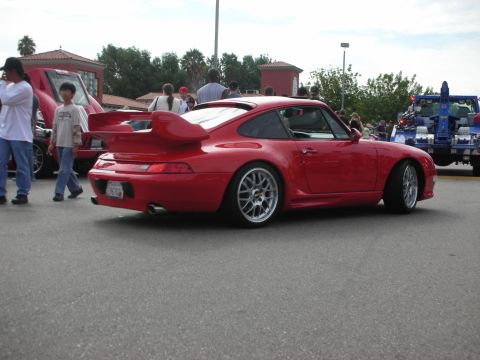 1998 Ford Crown Victoria - A red Porsche 911 with a prominent rear spoiler is parked on asphalt, with other vehicles and people in the background.