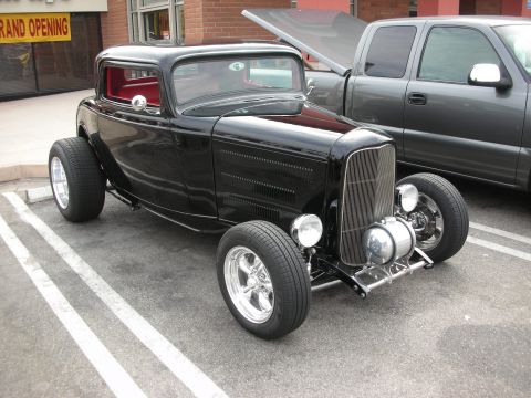 1998 Ford Crown Victoria - A classic black hot rod is parked in a lot next to a modern pickup truck with its hood open.