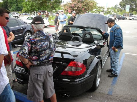 1998 Ford Crown Victoria - A group of people are gathered around a black convertible sports car in a parking lot.