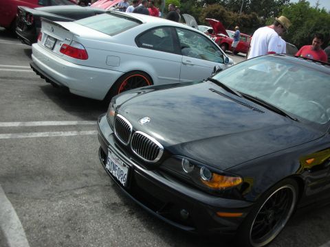 1998 Ford Crown Victoria - black BMW 3 Series coupe sits in the foreground with a silver BMW M3 on orange wheels behind it at a parking-lot meet.