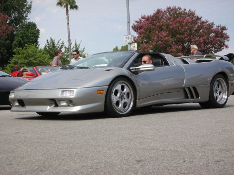 1998 Ford Crown Victoria - silver Lamborghini Diablo roadster with its top off sits in a parking lot, blooming trees and spectators behind.