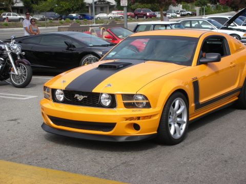 1998 Ford Crown Victoria - orange Ford Mustang with black racing stripes and a Saleen Parnelli Jones graphic parked in a lot beside a motorcycle.