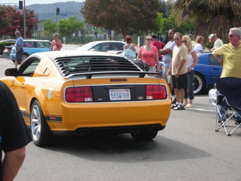 1998 Ford Crown Victoria - orange Ford Mustang Saleen with rear-window louvers and California plate seen from behind as spectators stand nearby.