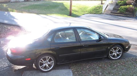1999 Ford Taurus - A black sedan is parked on a driveway, viewed from the side. The vehicle has aftermarket wheels and a body style consistent with early 2000…