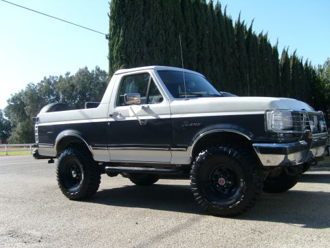 1989 Ford Bronco - Two-tone black and silver lifted Ford Bronco with brush guard and chrome wheels on mud tires, parked in front of cypress trees.