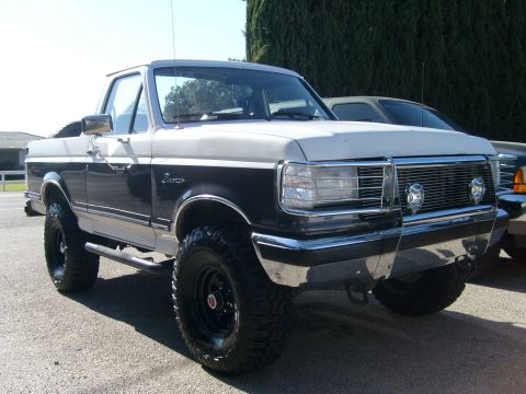 1989 Ford Bronco - Passenger-side front three-quarter view of a lifted two-tone dark gray and white Ford Bronco with its hardtop removed and roll bar exposed,…