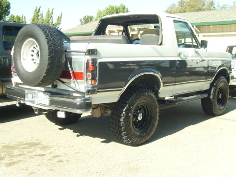 1989 Ford Bronco - Rear three-quarter view of a lifted dark-grey-over-white Ford Bronco with the rear top removed, showing the exposed rear bench and roll bar…