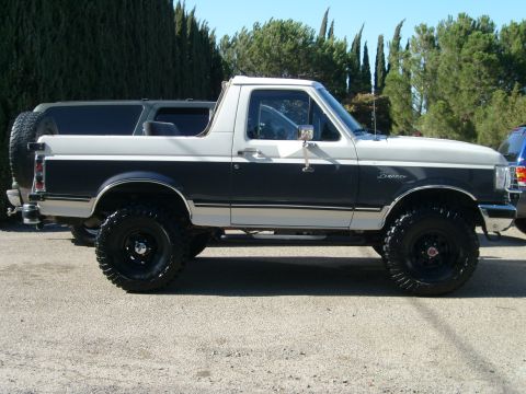 1989 Ford Bronco - Passenger-side profile of a lifted dark-grey-over-white Ford Bronco with the rear top removed, parked on gravel in front of cypress and pin…