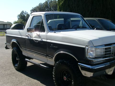 1989 Ford Bronco - Three-quarter front view of a lifted two-tone white-over-dark-blue Ford Bronco with its hardtop removed, parked on asphalt near trees and a…