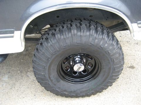 1989 Ford Bronco - Close-up of a rear wheel on a grey and white two-tone truck: a black steel wheel with a chrome center cap and an aggressive Big-O mud-terra…
