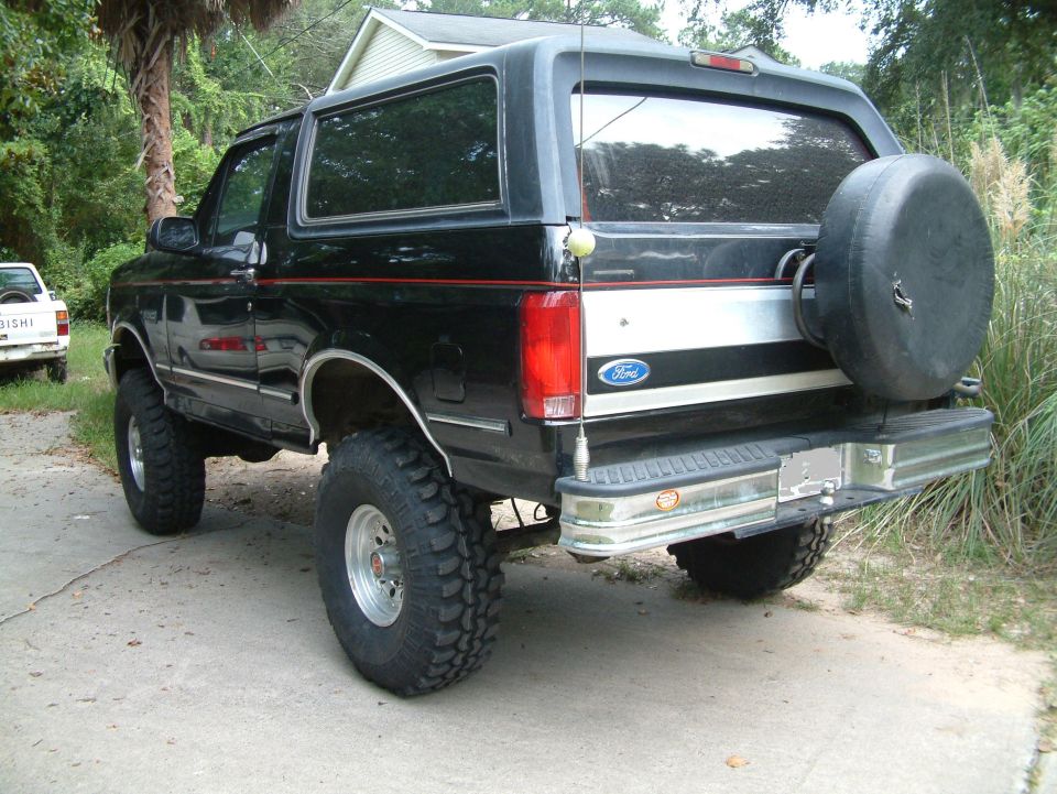 Black Ford Bronco with spare tire on back, parked on a driveway. Another white vehicle is in the background