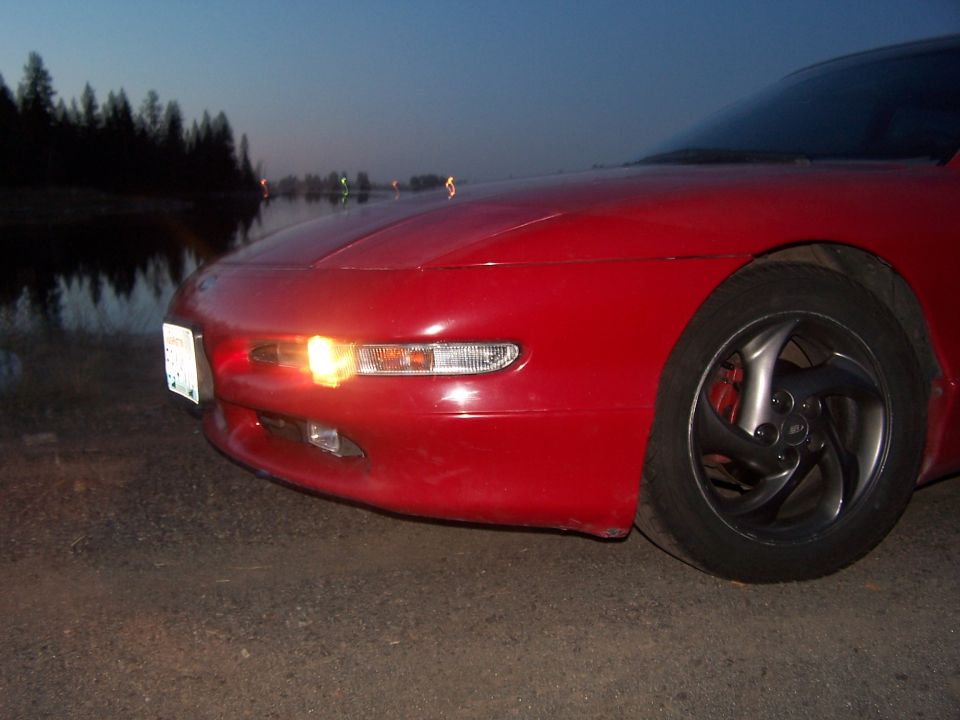 Red coupe front end at dusk by a lake, illuminated front light and dark alloy wheel