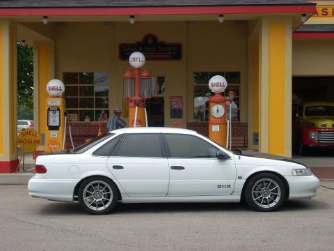 1992 Ford Taurus - Side view of a white 1992 Ford Taurus at a classic gas station with vintage pumps.
