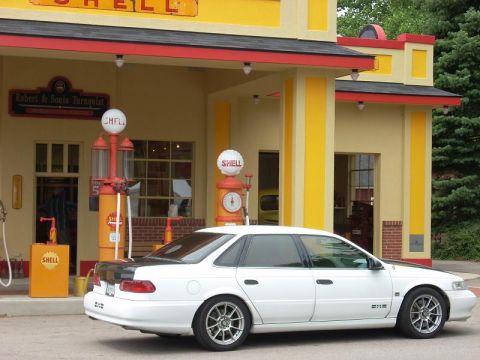 1992 Ford Taurus - A white sedan with aftermarket wheels is parked in front of a classic Shell gas station with vintage pumps.