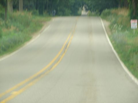 1992 Ford Taurus - A rural road ascends a hill, marked by double yellow lines and bordered by grass and trees. A sign is visible on the right side of the road.