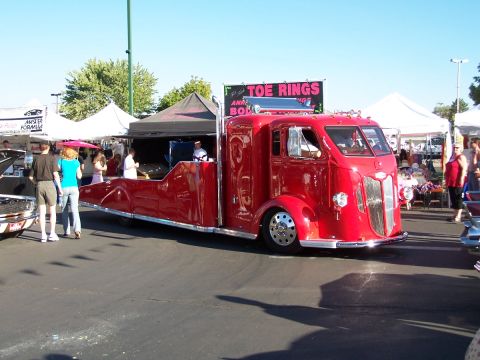 1990 Ford Bronco - A custom red truck with a flatbed trailer is displayed at an outdoor event with tents and people around.