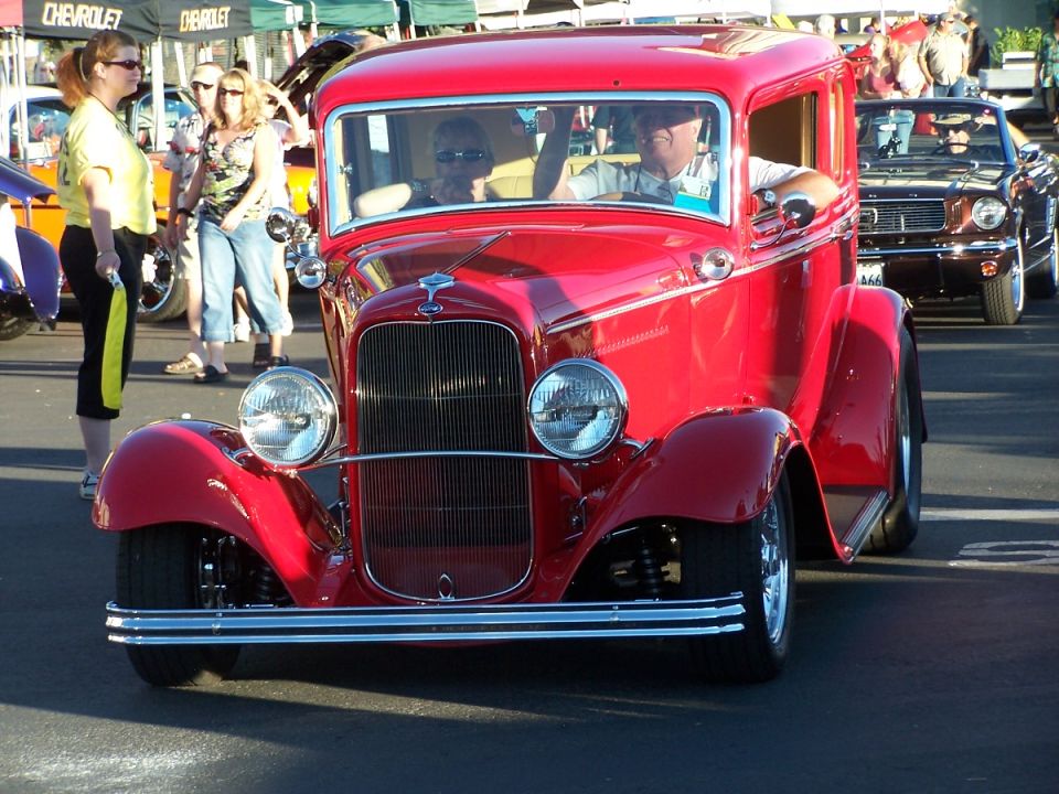 Red hot rod coupe with a chrome grille and two people inside. Another car is behind it