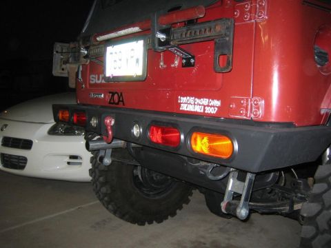 1987 Suzuki Samurai - The rear of a red off-road vehicle is shown, a custom bumper and a license plate. A white car is visible behind it.