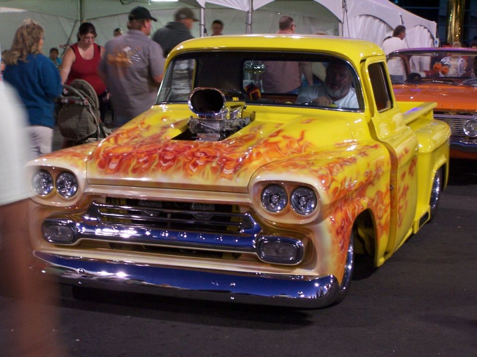 Yellow pickup truck with flame paint and a blower sticking out of the hood. Another truck is parked next to it
