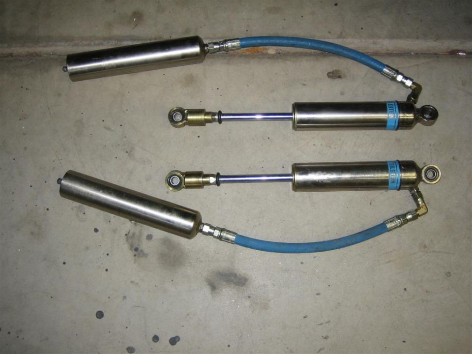Two silver shock absorbers with blue hoses and blue Bilstein labels on a concrete surface