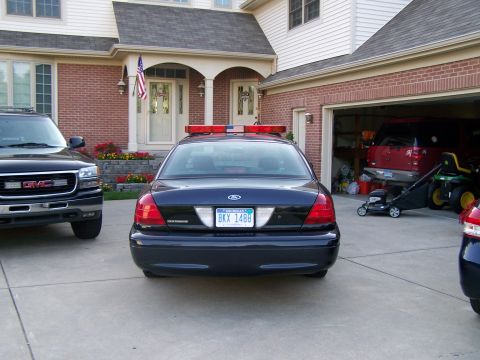 2003 Ford Crown Victoria - A police car and a GMC SUV are parked in a driveway in front of a house with an open garage.