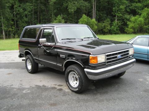 1995 Ford Bronco - This black Ford Bronco is parked on an asphalt surface with trees in the background.